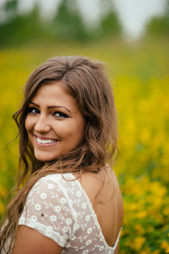 A young woman with long brown hair and a white lace dress smiles over her shoulder while standing in a field of yellow flowers, with greenery and blurred trees in the background. by Justin Salem Meyer