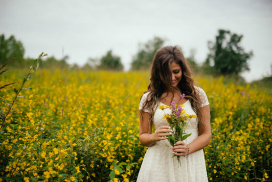 A woman in a white dress stands in a field of yellow wildflowers, holding a bouquet and smiling while looking down. The background is filled with greenery and a cloudy sky. by Justin Salem Meyer