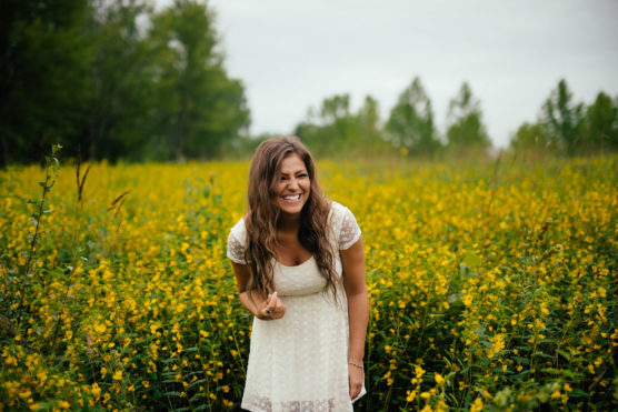 A woman with long brown hair in a white dress stands smiling and laughing in a field of tall yellow wildflowers, with green trees and a cloudy sky in the background. by Justin Salem Meyer
