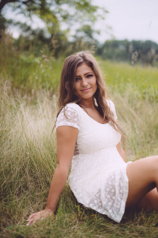 A woman with long brown hair wearing a white lace dress sits on the grass in a meadow, looking at the camera with a gentle smile. Trees and greenery are visible in the background. by Justin Salem Meyer