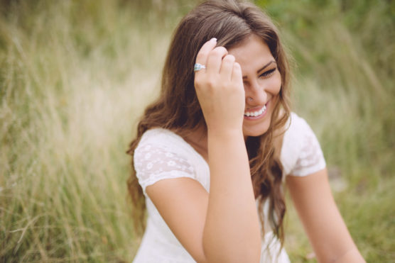 A young woman with long brown hair and a white lace dress sits outdoors on grass, smiling brightly and holding her hand near her face. by Justin Salem Meyer