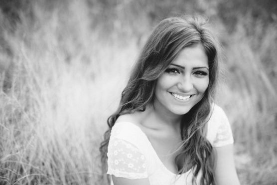 A young woman with long hair sits in tall grass, smiling warmly at the camera. She wears a light-colored, short-sleeved top and has a nose piercing. The photo is in black and white. by Justin Salem Meyer