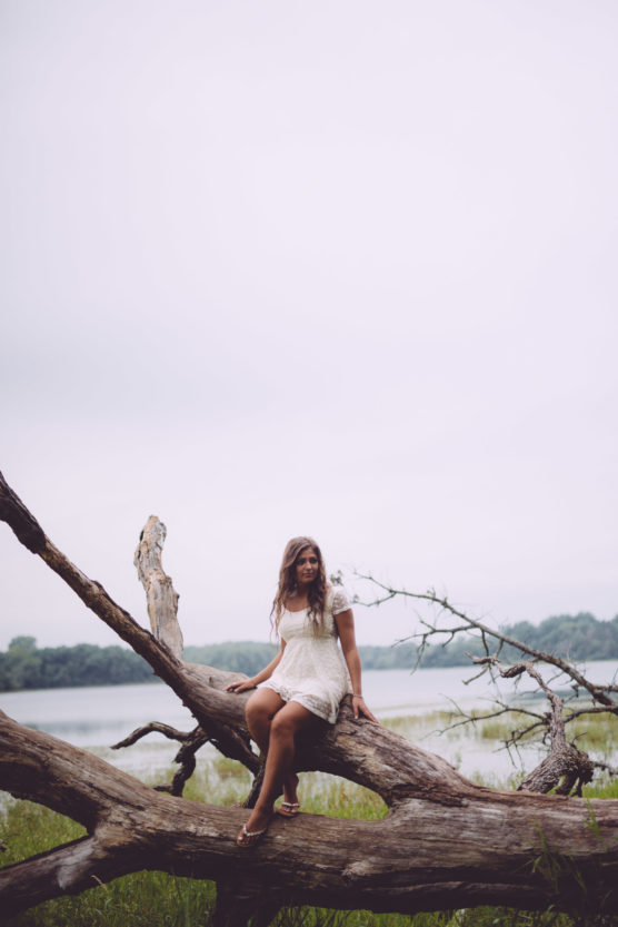 A woman in a short white dress sits on a large fallen tree by a lake, surrounded by green grass and trees under an overcast sky. by Justin Salem Meyer