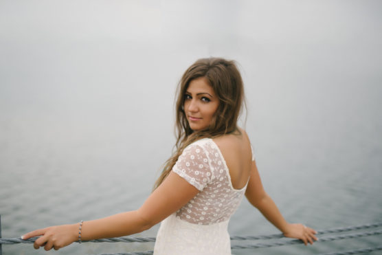 A woman in a white, short-sleeved dress with lace detail stands by a railing, looking over her shoulder toward the camera, with a calm, foggy body of water in the background. by Justin Salem Meyer