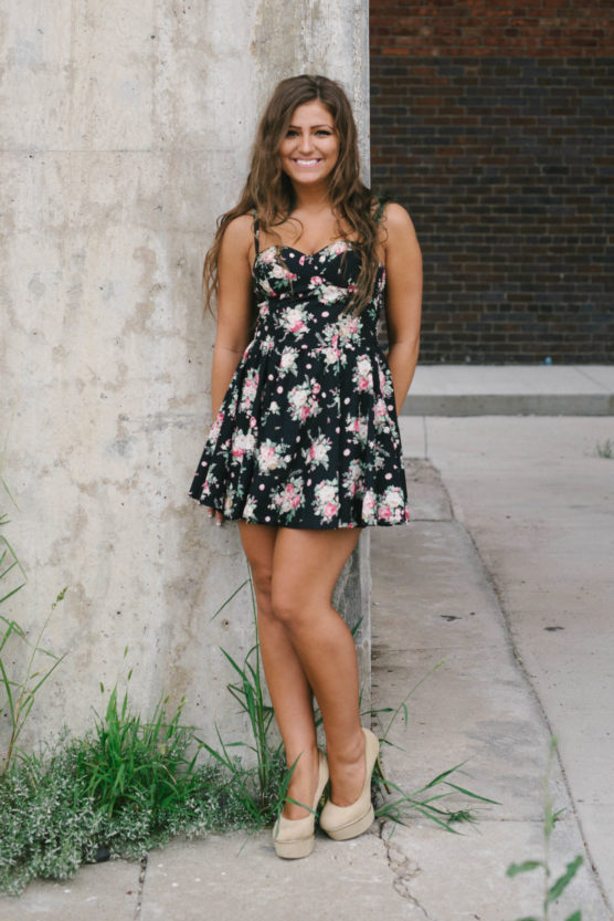 A woman with long brown hair in a black floral dress and beige shoes stands smiling against a tall concrete pillar, with grass at her feet and a brick wall in the background. by Justin Salem Meyer