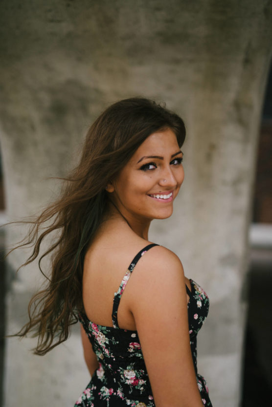 A young woman with long brown hair, wearing a black floral dress, stands sideways and smiles at the camera. The background is blurred concrete, and her hair is gently blowing in the wind. by Justin Salem Meyer