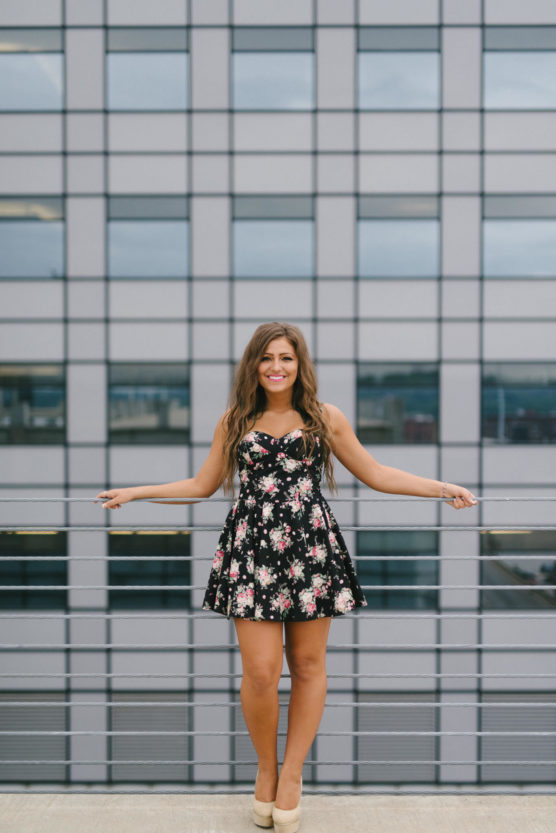 A woman in a black floral dress and beige heels stands smiling on a rooftop, holding onto a railing, with a modern glass building in the background. by Justin Salem Meyer