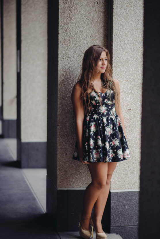 A young woman with long hair, wearing a sleeveless black floral dress and beige high heels, stands leaning against a textured wall in a corridor, looking thoughtfully into the distance. by Justin Salem Meyer