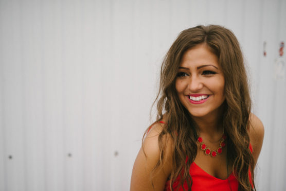 A young woman with long brown hair and a bright smile wears a red dress and matching red necklace, standing in front of a white corrugated metal wall. by Justin Salem Meyer