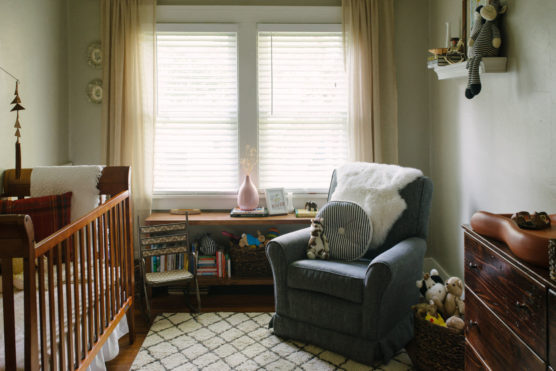Cozy nursery with a wooden crib, a cushioned armchair with a striped pillow and stuffed toy, a dresser with plush animals, a patterned rug, and a bookshelf, all softly lit by natural light from two large windows. by Justin Salem Meyer