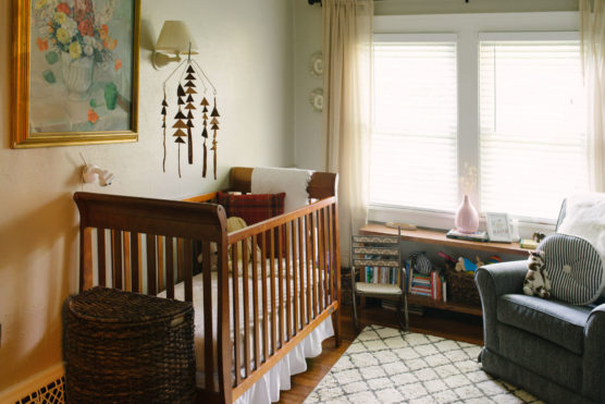 A cozy nursery with a wooden crib, wicker hamper, wall art, hanging mobile, armchair, bookshelf with toys and books, and sunlight streaming through large windows with light curtains. by Justin Salem Meyer