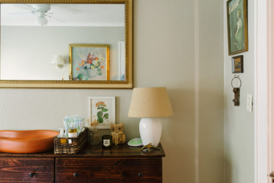 A wooden dresser with a lamp, framed artwork, basket of baby supplies, and small decor sits below a large mirror reflecting a painting of flowers in a vase on a pale wall. Vintage keys hang on the adjacent wall. by Justin Salem Meyer