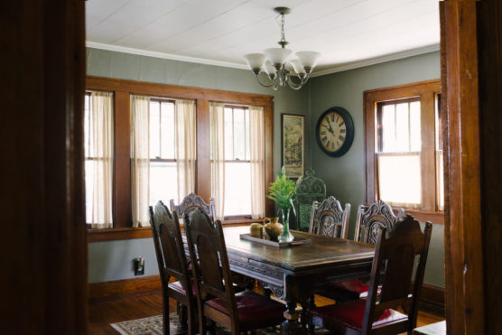A cozy dining room with a wooden table, six ornate chairs, a chandelier, large windows with sheer curtains, a wall clock, and green plants as centerpiece decor. Warm sunlight fills the space. by Justin Salem Meyer