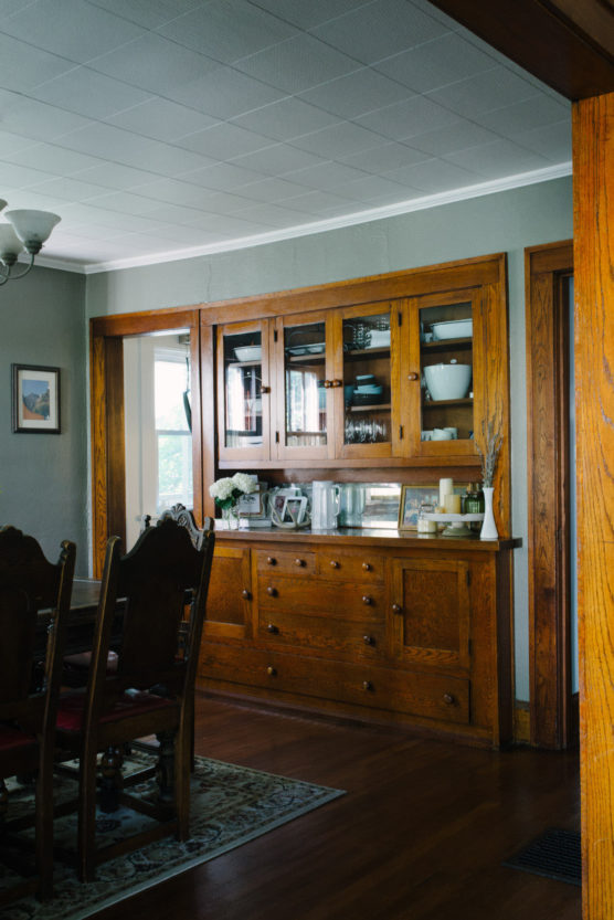 A wooden dining room hutch with glass doors displays dishes and decor. Antique wooden chairs and a dining table sit on a patterned rug. Light streams in from a nearby window. by Justin Salem Meyer