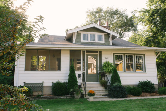 A white bungalow-style house with a small porch, green door, large windows, and neatly trimmed shrubs and plants in the front yard, set against a backdrop of trees on a sunny day. by Justin Salem Meyer