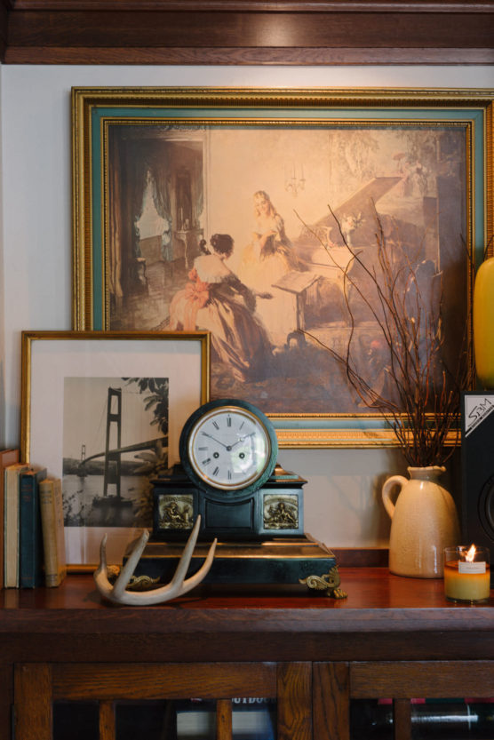 A mantel displays a framed painting of women at a piano, a black-and-white photo of a bridge, a clock, books, antlers, a ceramic jug with twigs, and a lit candle. by Justin Salem Meyer