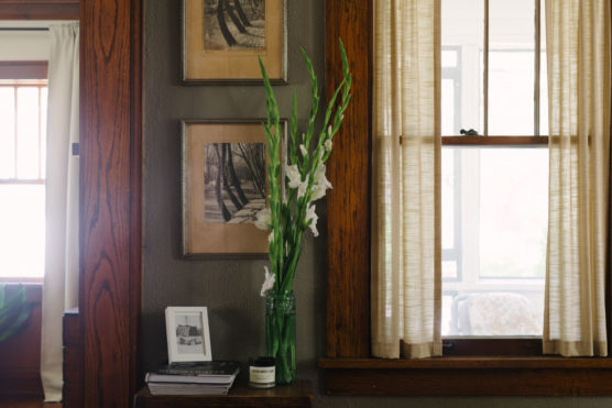 A vase with tall white flowers sits on a wooden table beside a window with sheer curtains. Framed black-and-white photos hang above, and a small photo frame, a candle, and books rest on the table. by Justin Salem Meyer