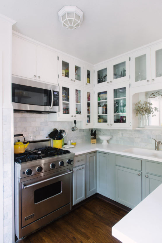 A bright, modern kitchen with white upper cabinets featuring glass doors, light blue lower cabinets, stainless steel stove and microwave, white countertops, and wooden floor. Yellow cookware sits on the stove. by Justin Salem Meyer
