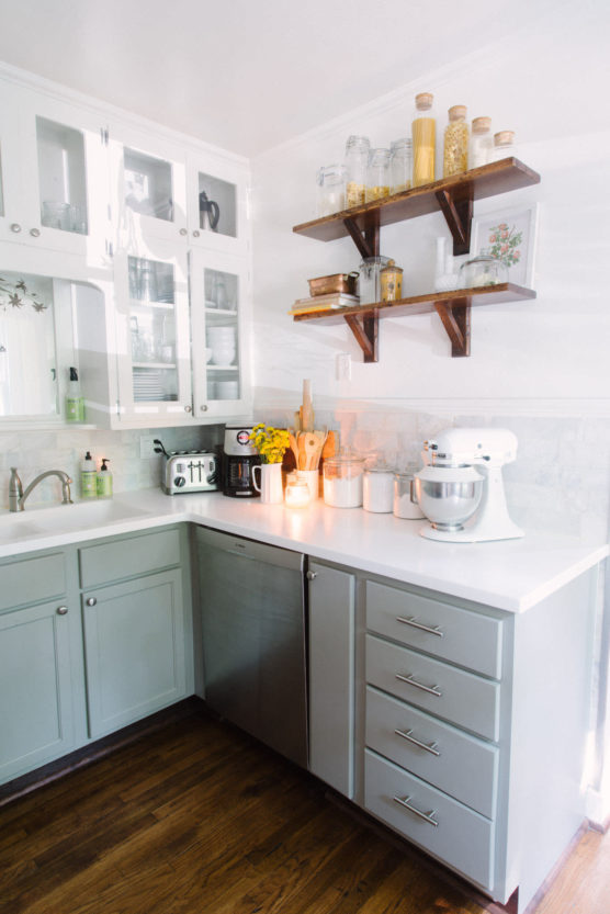 Bright kitchen with light green cabinets, white countertops, a stand mixer, toaster, and jars on open shelves. Sunflowers are in a vase by the sink, and the room features wood floors and glass-front cabinets. by Justin Salem Meyer