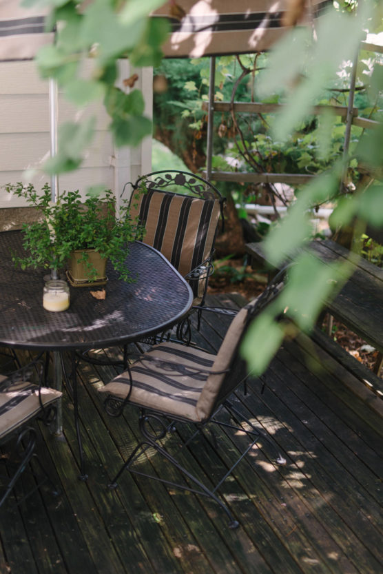 A cozy outdoor patio with a black metal table and striped cushioned chairs, a potted plant, and candles, surrounded by greenery and dappled sunlight. by Justin Salem Meyer