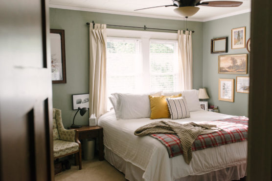 A cozy bedroom with a neatly made bed, white bedding, and yellow and striped pillows. A plaid blanket and knit throw are at the foot of the bed. Light streams through a window with cream curtains. Artwork decorates the walls. by Justin Salem Meyer