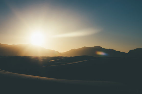 Sunrise over a desert landscape with smooth sand dunes in the foreground and mountains in the background, bathed in warm golden light. by Justin Salem Meyer