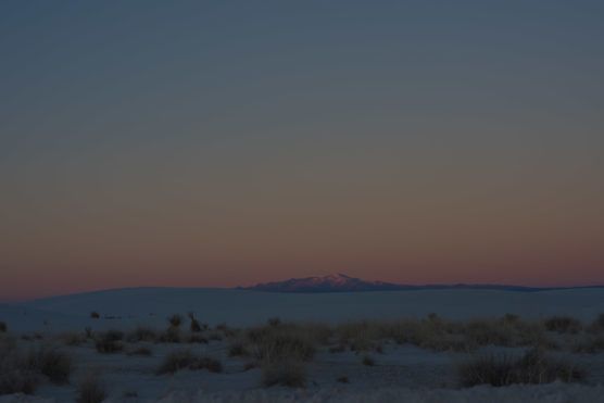 Distant snow-capped mountains under a dusky sky with soft pink and purple hues, seen from a desert landscape with sparse bushes and sand in the foreground. by Justin Salem Meyer