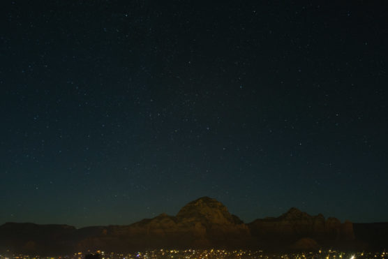 A clear night sky filled with stars above dark, rocky mountains; a small town with scattered lights glows at the base of the mountains. by Justin Salem Meyer