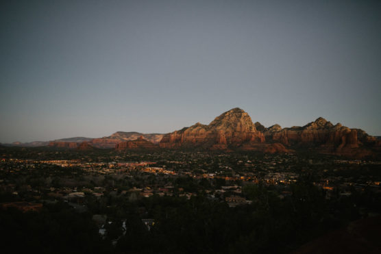 A panoramic view of red rock mountains at dusk, with a town nestled at their base and the sky gradually darkening above. by Justin Salem Meyer