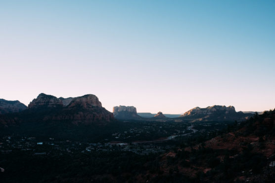 Wide view of a desert landscape at sunset, featuring rocky red mountains and buttes under a clear blue sky, with sparse vegetation and scattered buildings in the foreground. by Justin Salem Meyer