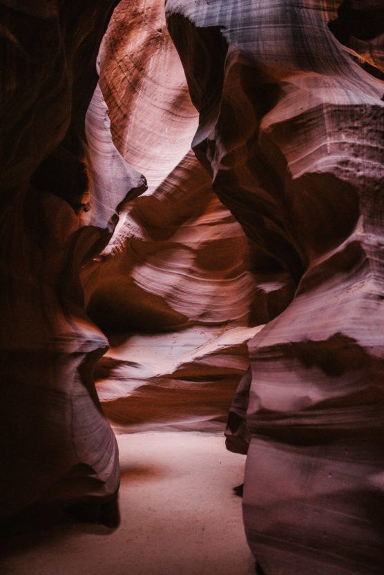Sunlight filters into a narrow slot canyon, illuminating smooth, wave-like sandstone walls in shades of red and orange, creating dramatic light and shadow patterns along the sandy canyon floor. by Justin Salem Meyer