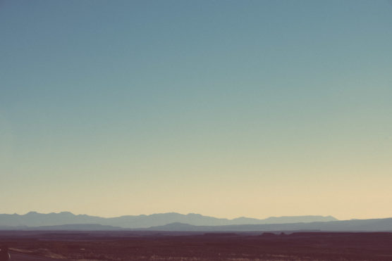 A wide, hazy desert landscape with distant mountain ranges under a clear sky, bathed in soft, muted sunlight. The scene has a tranquil, minimalist feel. by Justin Salem Meyer