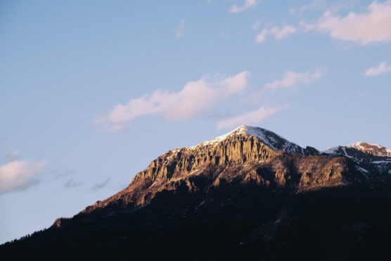 Sunlit mountain peak with patches of snow, rugged cliffs, and a clear blue sky with a few scattered clouds at sunset. Shadows cover the lower part of the mountainside. by Justin Salem Meyer