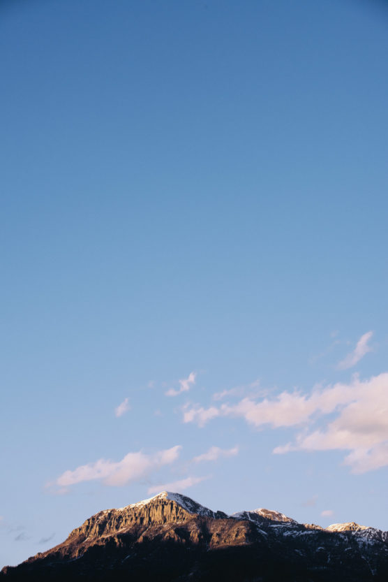 Snow-capped mountain peak with rocky slopes, bathed in warm sunlight, under a vast blue sky with a few scattered clouds. by Justin Salem Meyer