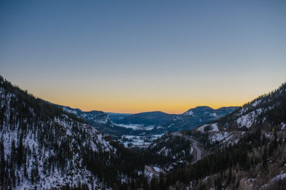 A winding road cuts through a snowy mountain valley at dusk, with forested slopes on either side and a golden-yellow sunset glowing above distant peaks. by Justin Salem Meyer