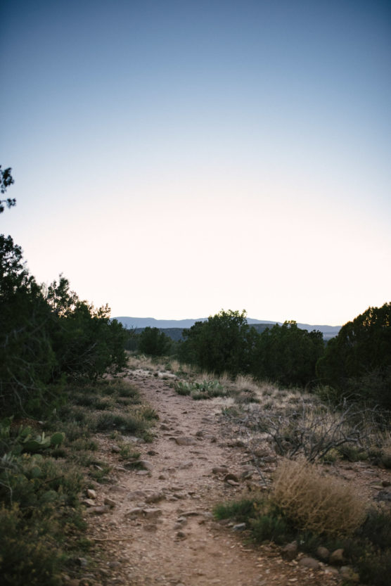 A rocky dirt trail winds through dry grass and bushes with scattered green shrubs, leading toward distant mountains under a clear blue sky at sunset. by Justin Salem Meyer