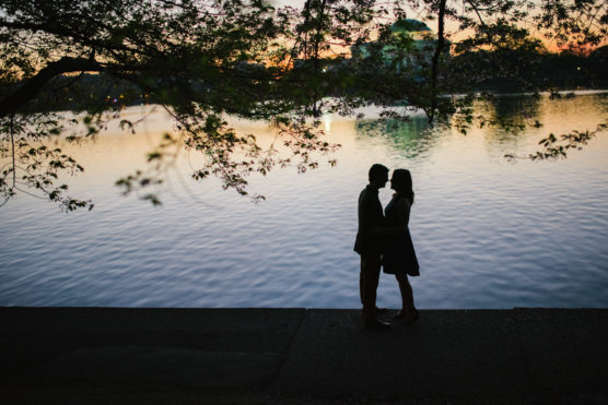 Silhouette of a couple standing close together by a calm lakeside at sunset, framed by overhanging tree branches with soft light reflecting on the water. by Justin Salem Meyer