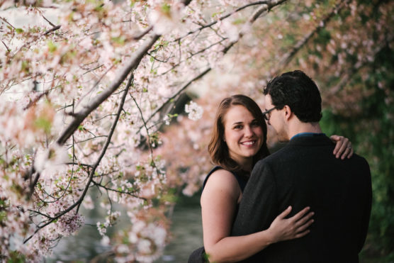 A woman smiles at the camera while embracing a man under blooming cherry blossom trees. The background features soft pink flowers and a blurred river, creating a romantic springtime scene. by Justin Salem Meyer