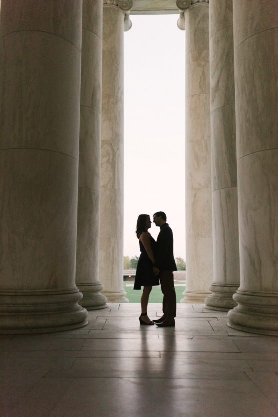 A couple stands facing each other and holding hands, silhouetted between large marble columns with soft daylight in the background. by Justin Salem Meyer