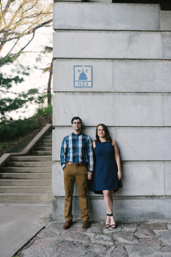 A man and woman hold hands while standing against a stone wall near outdoor steps. The man wears a plaid shirt and khaki pants; the woman wears a blue dress and sandals. Above them is a blue D.C. marker. by Justin Salem Meyer