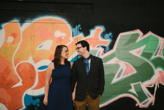 A woman in a blue dress and a man in a blazer and glasses stand holding hands, smiling at each other, in front of a colorful graffiti-covered wall. by Justin Salem Meyer