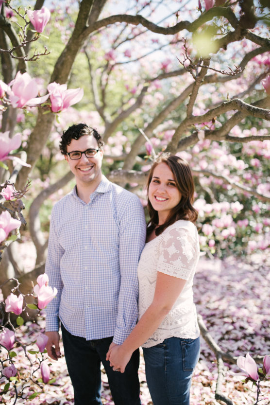 A man and a woman stand holding hands and smiling beneath blooming pink magnolia trees, with sunlight filtering through the branches. The ground is covered with fallen petals. by Justin Salem Meyer