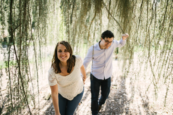 A smiling woman leads a man by the hand through hanging tree branches in a sunlit, wooded area. Both appear happy and relaxed, enjoying the natural surroundings. by Justin Salem Meyer