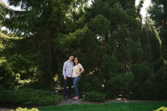 A couple stands closely together and smiles in front of tall, lush pine trees in a sunny outdoor park or garden. by Justin Salem Meyer