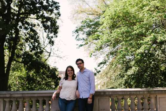 A woman and a man stand together, smiling, on a stone bridge surrounded by lush green trees on a sunny day. by Justin Salem Meyer