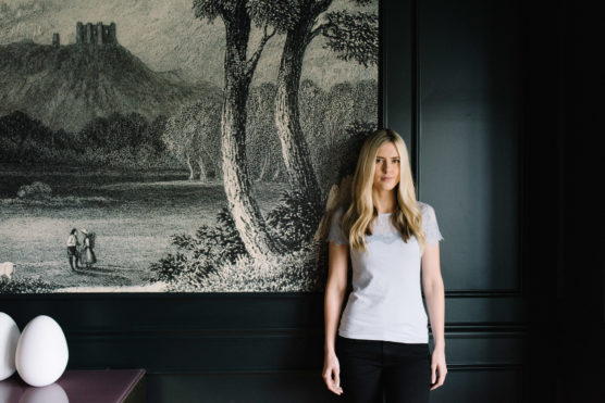A woman with long blonde hair stands indoors against a wall featuring a detailed black-and-white mural of trees and distant ruins. She wears a short-sleeved light gray top and black pants. by Justin Salem Meyer