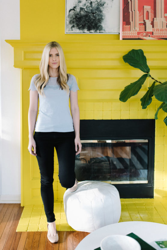 A blonde woman stands in front of a bright yellow fireplace with one foot resting on a white pouf. She wears a light gray t-shirt, black pants, and flats. A green plant and framed art decorate the cozy, modern room. by Justin Salem Meyer