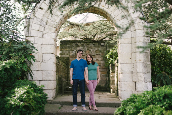 A man and a woman stand holding hands under a stone archway in a lush garden, smiling at the camera. The scene is framed by greenery and old stone walls. by Justin Salem Meyer