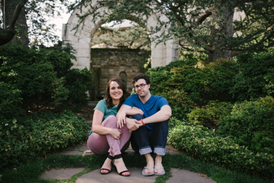 A smiling woman and a man sit side by side on a stone path surrounded by green bushes, with an old stone archway in the background. The woman wears a teal shirt and pink pants; the man wears glasses, a blue shirt, and jeans. by Justin Salem Meyer