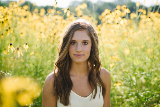 A young woman with long brown hair stands in a field of yellow wildflowers, looking at the camera. She is wearing a light yellow tank top and the sun is shining, creating a soft, natural background. by Justin Salem Meyer